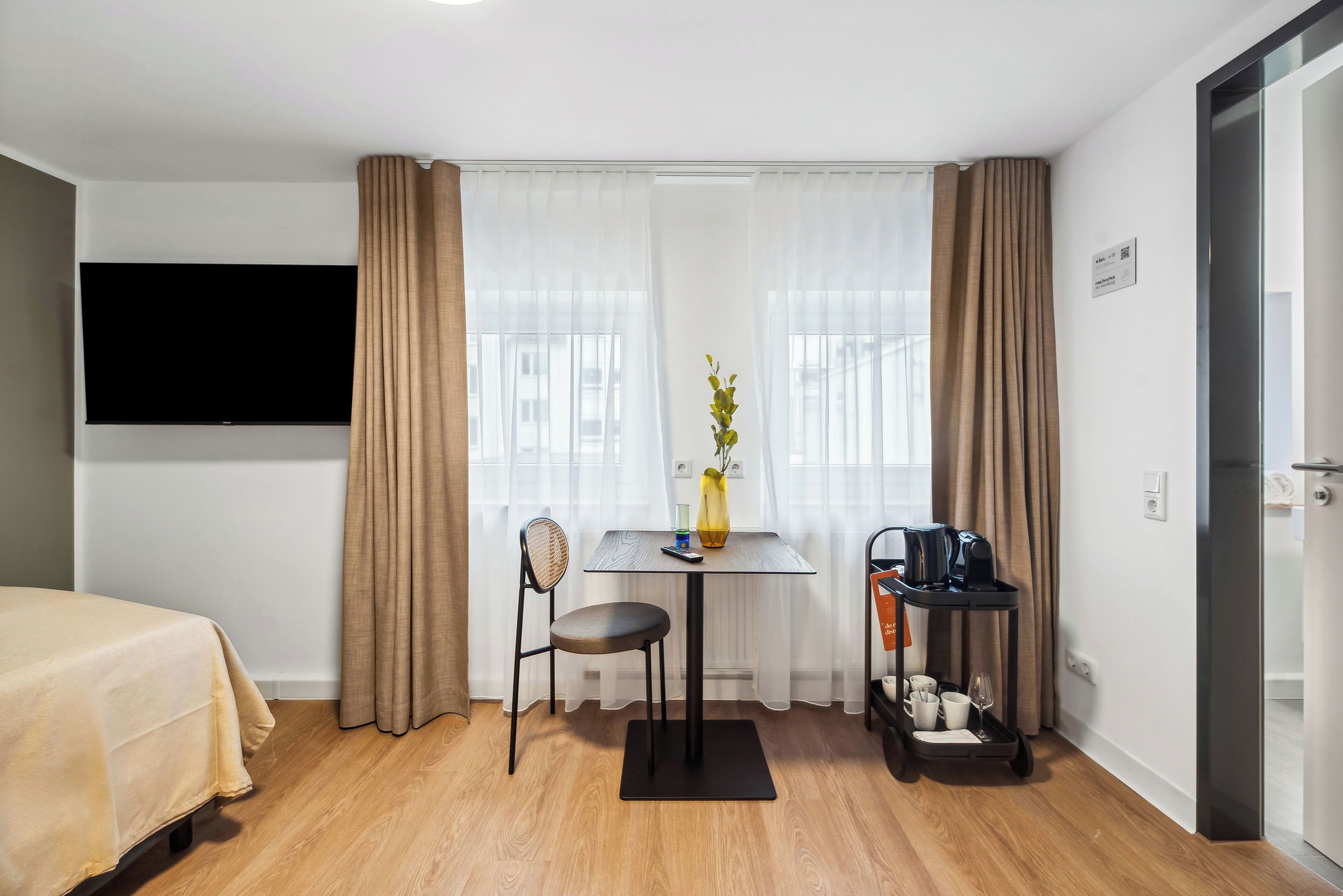 Habitación de hotel con cama, televisor, una mesita con un florero y una silla, además de una cafetera.