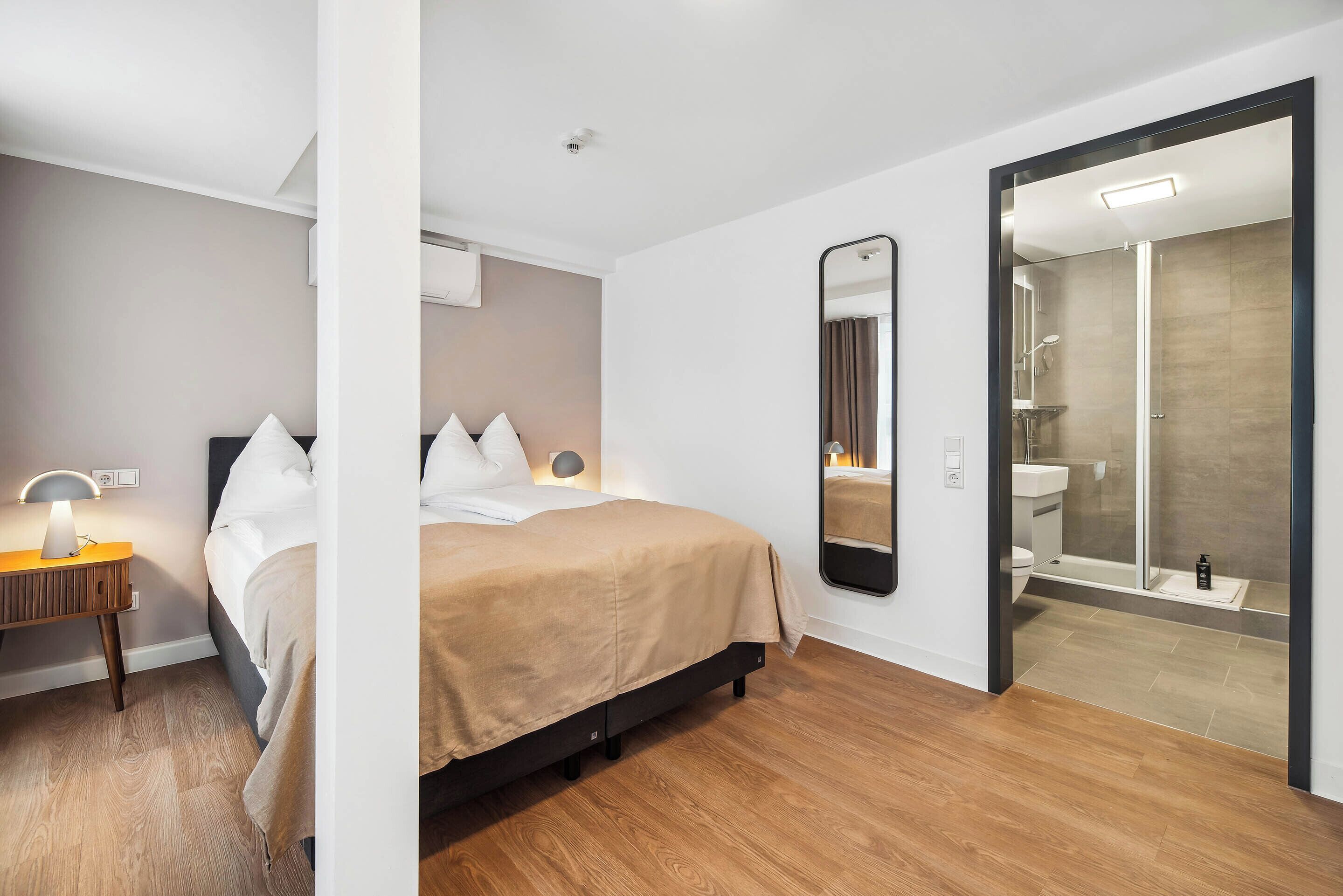 Una habitación de hotel moderna con cama, mesita de noche, lámpara, espejo y zona de ducha.