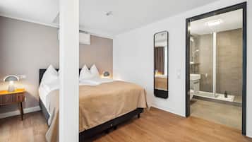 Una habitación de hotel moderna con cama, mesita de noche, lámpara, espejo y zona de ducha.