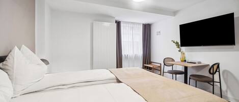 A hotel room with a bed, a desk, two chairs, a TV, and a window with curtains.