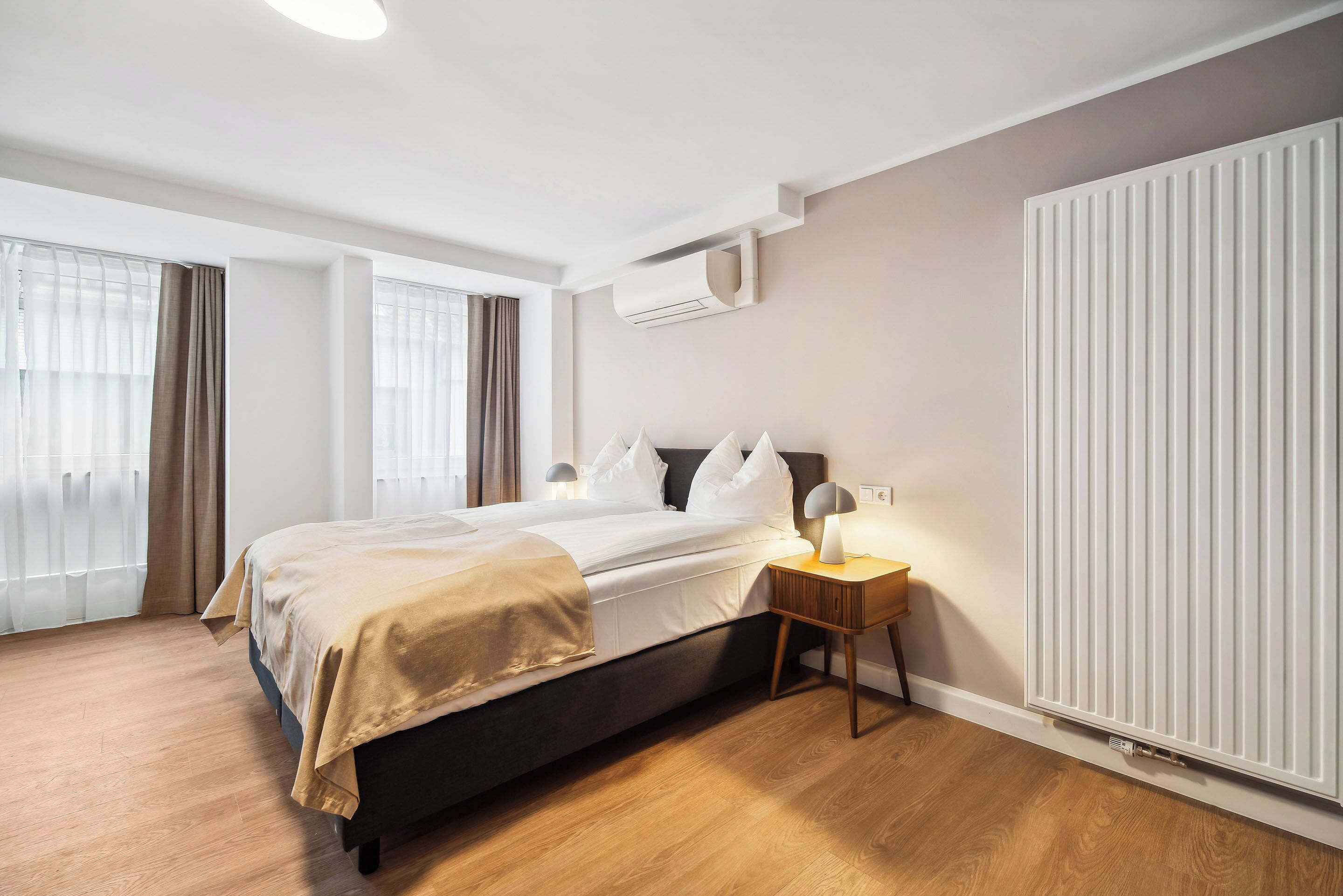 Una habitación de hotel moderna con una cama grande, lámparas de noche, un radiador y una ventana con cortinas.