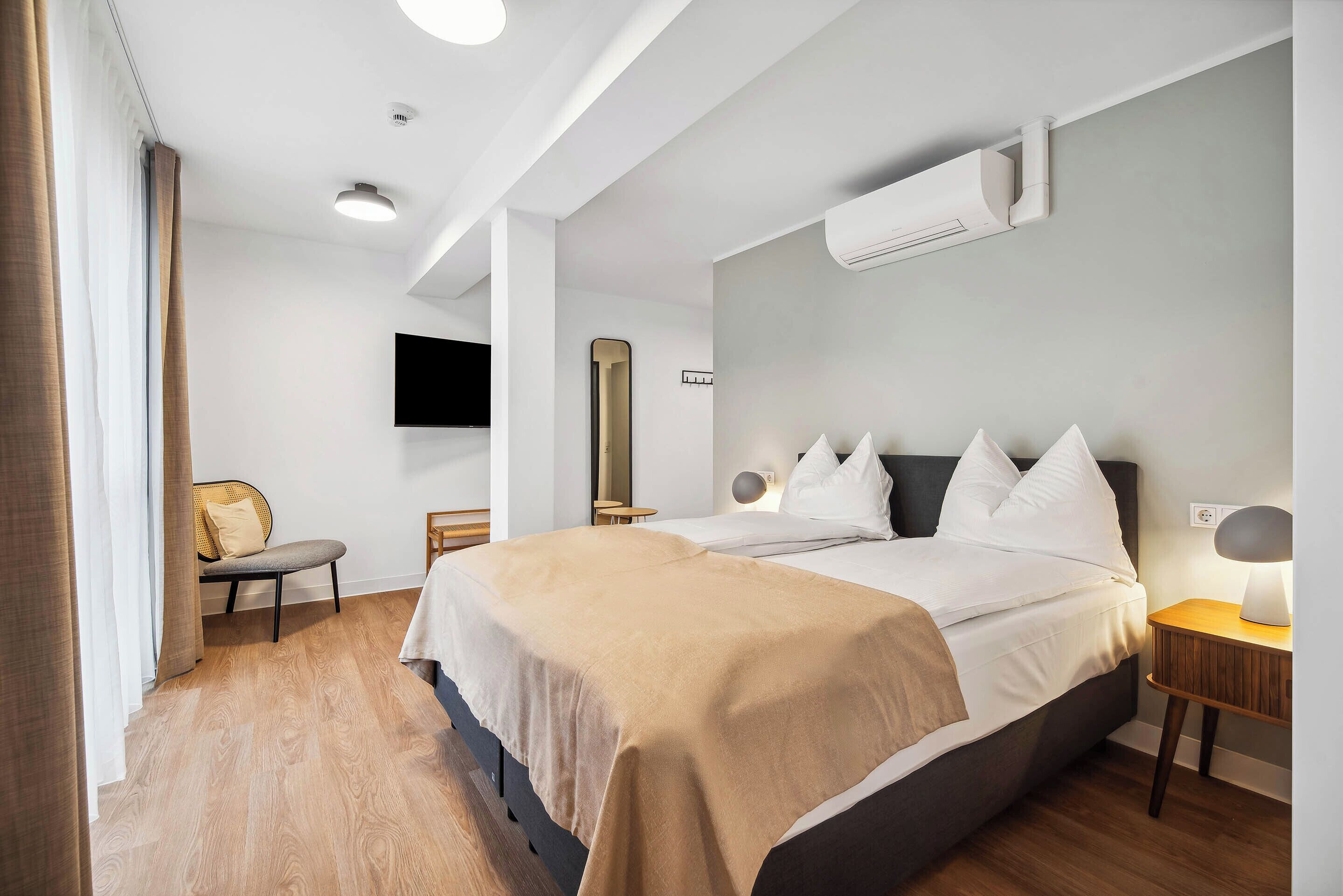 Habitación de hotel moderna con una cama grande, una mesita de noche con lámpara, un televisor de pantalla plana en la pared y una ventana con cortinas.