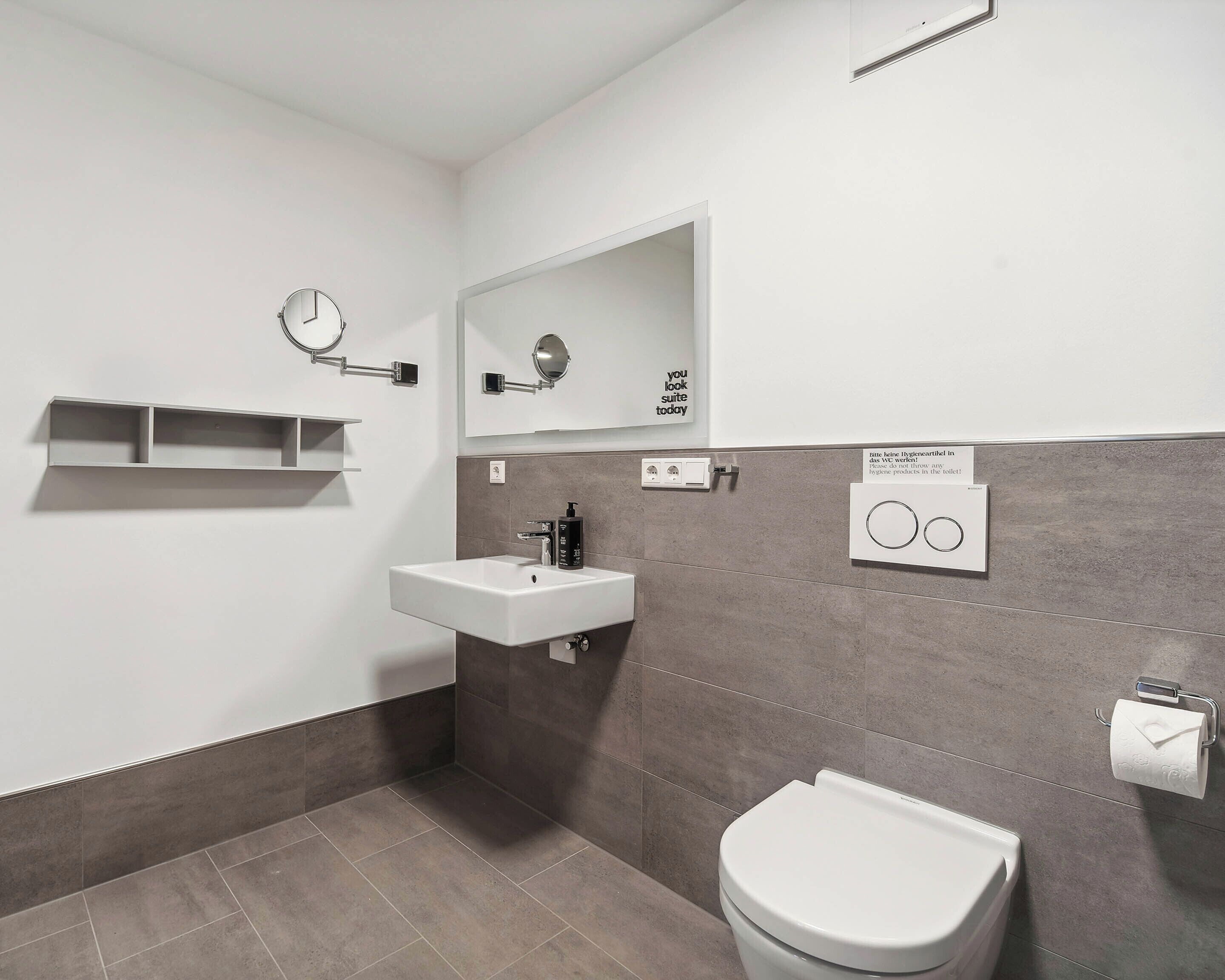 Een moderne badkamer met een wit wastafelmeubel, een spiegel, toilet en een handdoekdispenser.