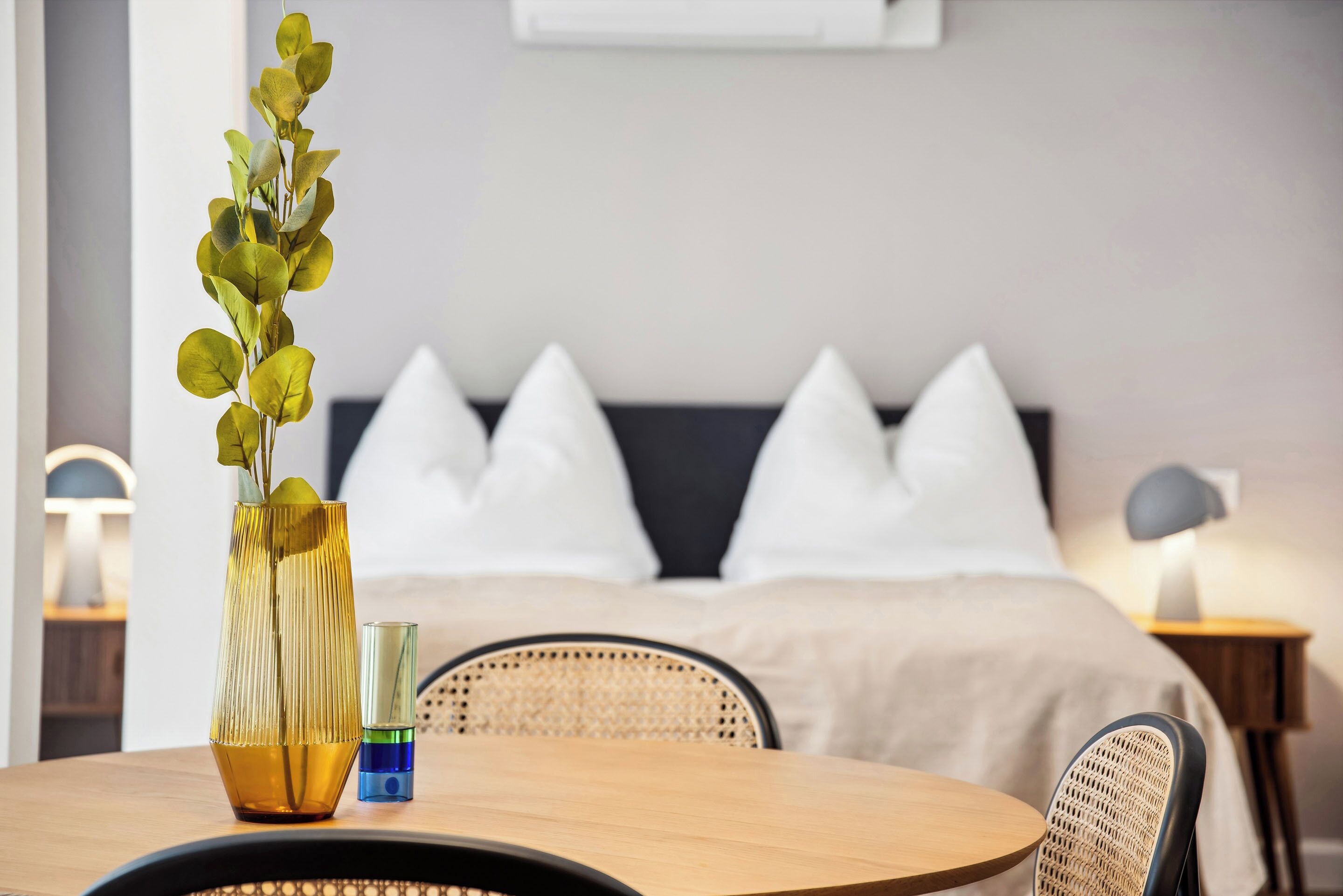 Ein modernes Hotelzimmer mit einem Holztisch, einer Vase mit grünen Blättern, einem Nachttisch mit Lampe und einem Bett mit weißen Kissen.