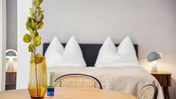 Ein modernes Hotelzimmer mit einem Holztisch, einer Vase mit grünen Blättern, einem Nachttisch mit Lampe und einem Bett mit weißen Kissen.