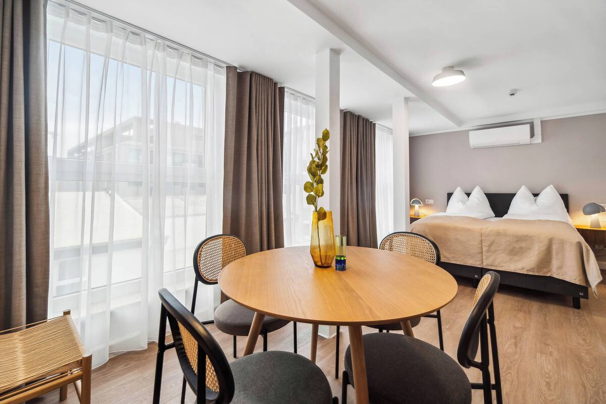 Une chambre d’hôtel avec une table ronde en bois, deux chaises, un lit avec des oreillers blancs et une grande fenêtre ornée de rideaux légers.