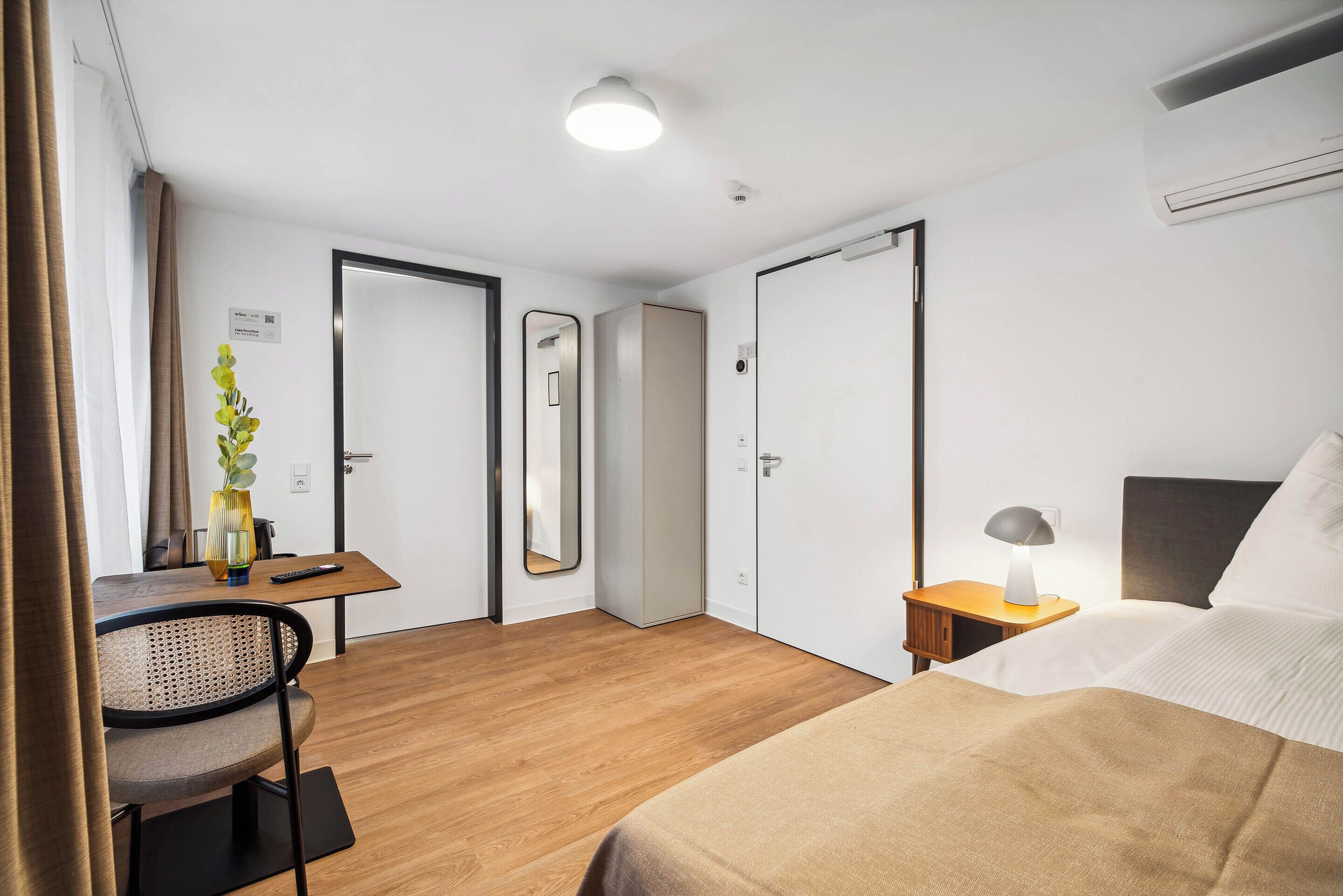 A hotel room with a bed, a desk with a chair, a small table with a lamp, and a mirror.