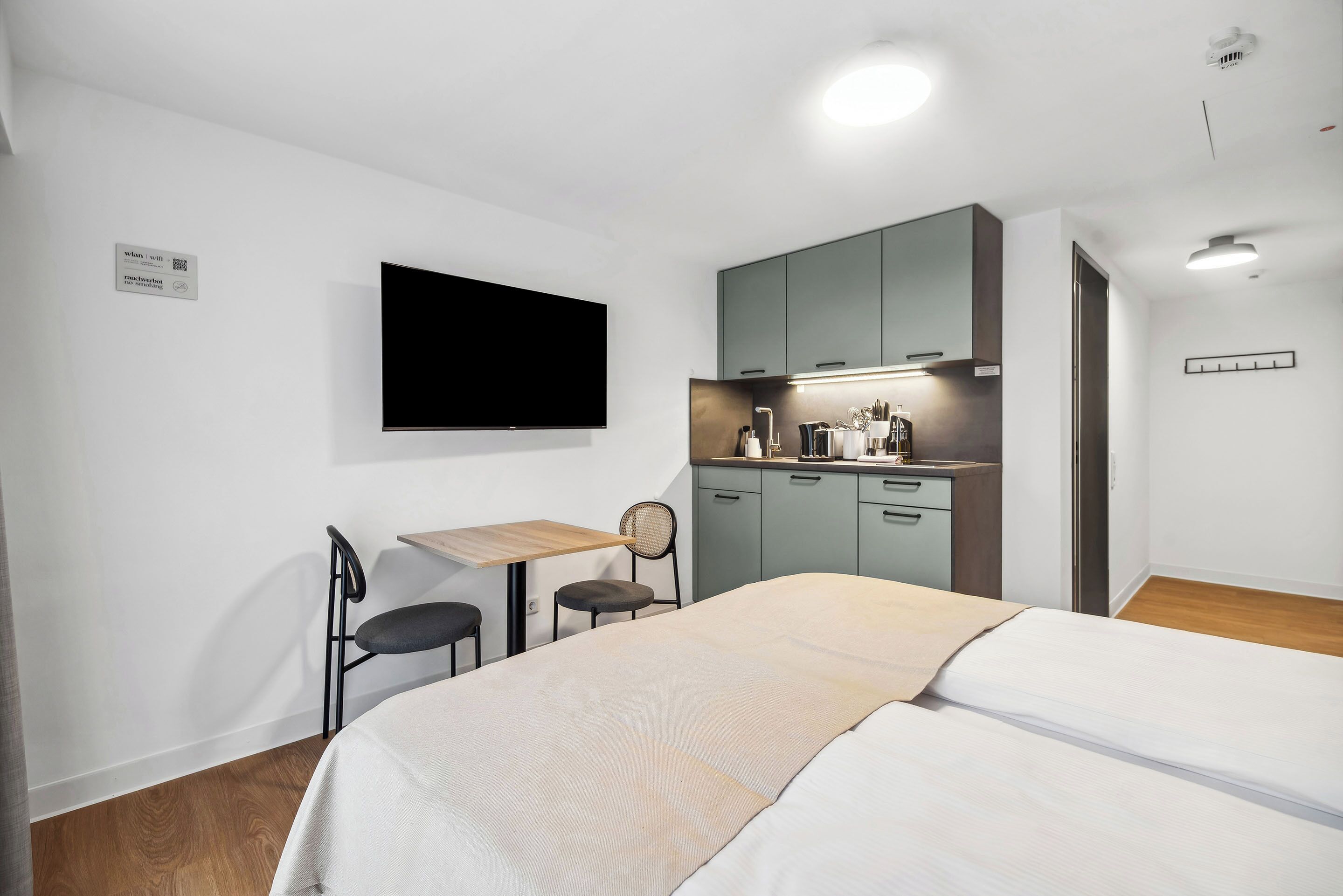Ein modernes Hotelzimmer mit Bett, Schreibtisch, Stuhl, Fernseher und einer Küchenzeile.
