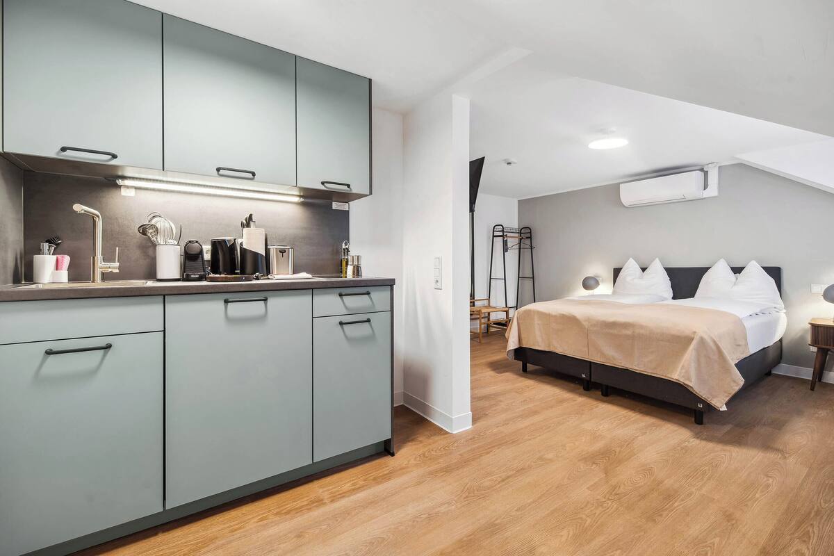 Une cuisine moderne avec des meubles gris et un plancher en bois, qui donne sur une chambre à coucher équipée d’un lit et d’une table de chevet.