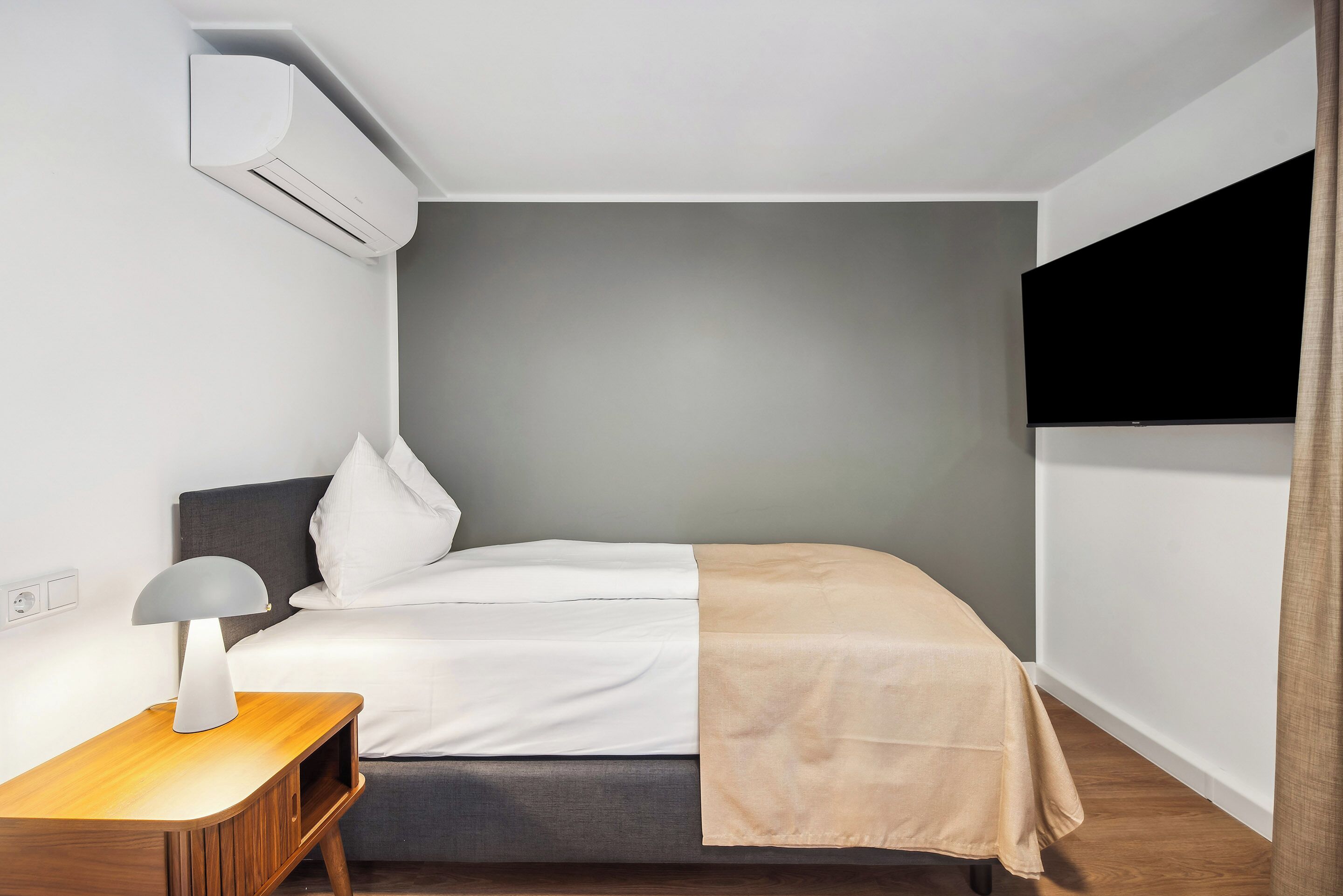 Una camera d'albergo moderna con un letto, un comodino con una lampada, una TV a muro e un condizionatore d'aria.