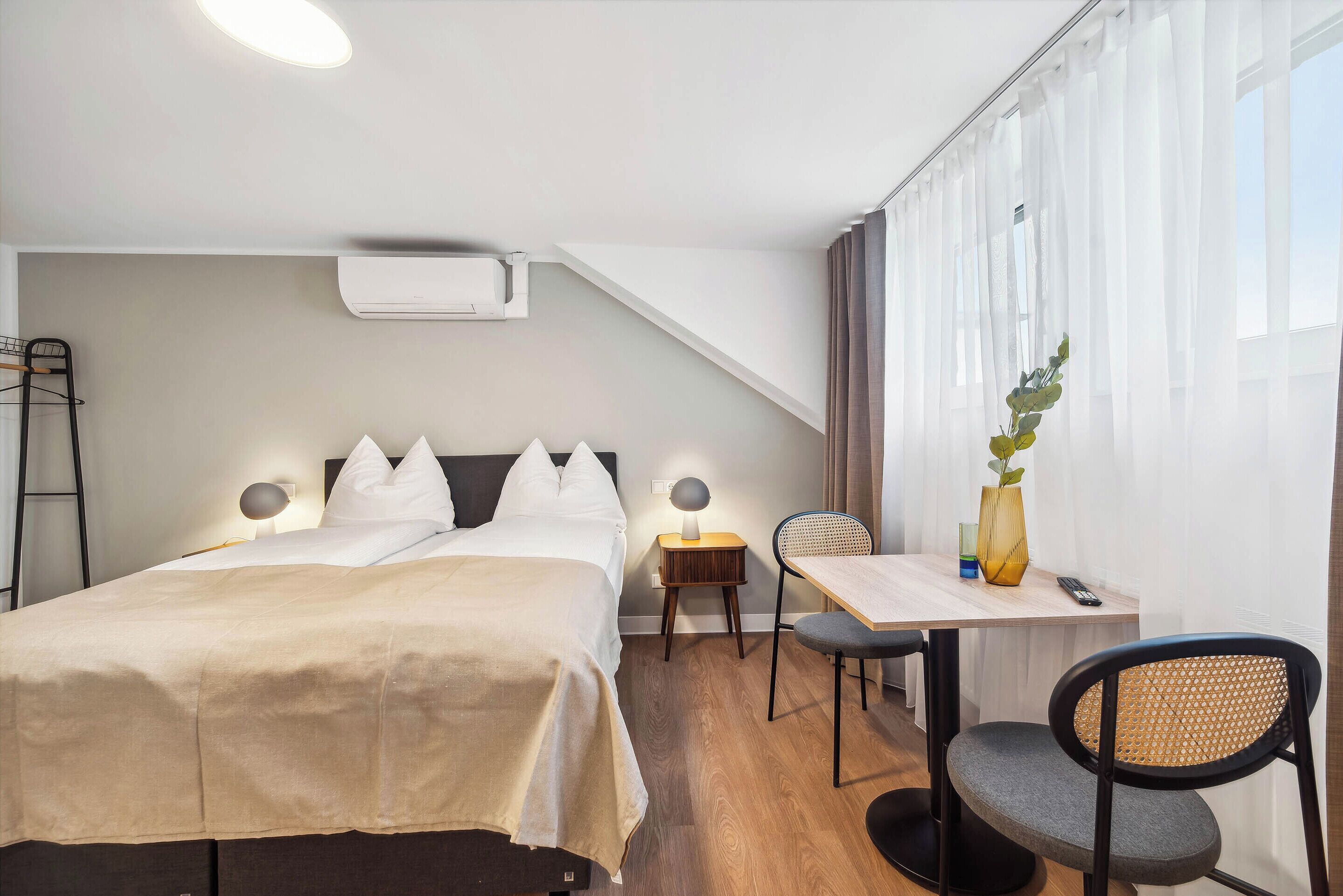 A hotel room with a bed, bedside table, lamp, chair, and a small table with a vase and remote control.