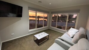 A modern living room with a flat-screen TV mounted on the wall, a seating area with a sofa and armchairs, a coffee table, and large windows offering a view of a parking lot and trees. - Modern waterfront luxury escape near Shepparton (Mooroopna)