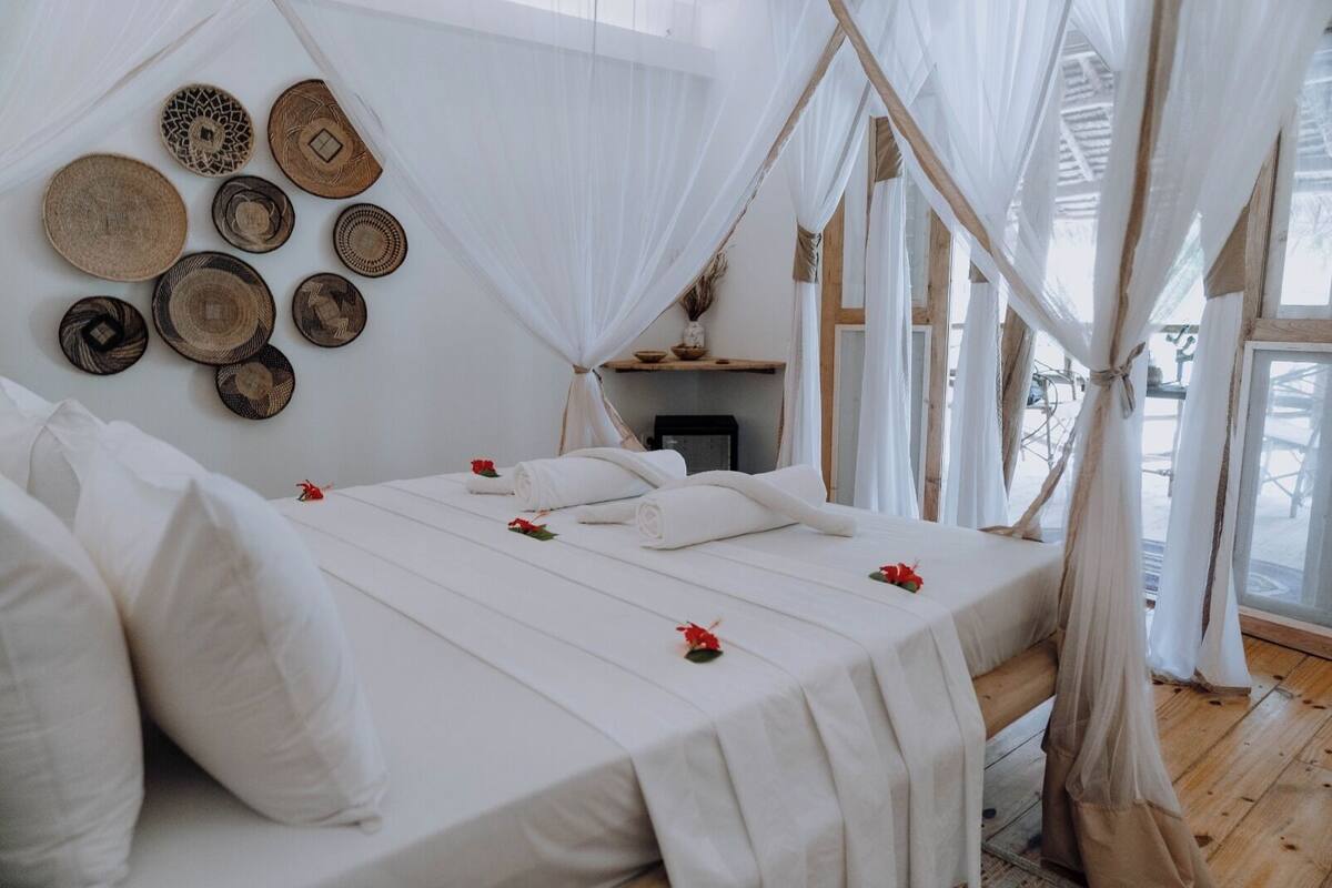 Ein Zimmer mit einem Bett, weißer Bettwäsche, einem Baldachin und an der Wand befestigten gewebten Körben.