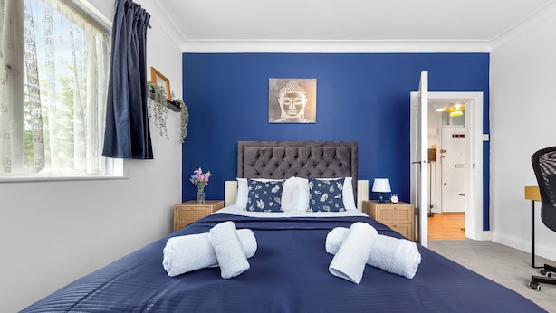 A neatly made bed with two towels, a headboard, and a wall-mounted Buddha artwork in a room with blue walls. - Amazing 3-bed Flat in Golders Green Near Station (London)