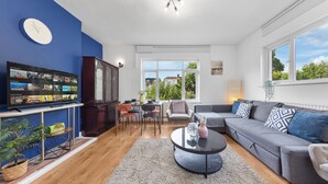 A modern living room with a blue accent wall, a flat-screen TV, a grey sofa, and a dining area with chairs and a table. - Amazing 3-bed Flat in Golders Green Near Station (London)