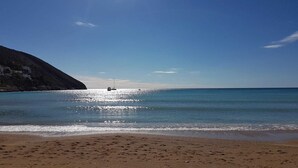 Une plage à l’eau d’un bleu cristallin, un bateau à l’horizon et une montagne en arrière-plan.