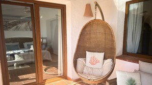 Une chaise en osier avec un coussin et un oreiller décoratif, un canapé avec un coussin, une fenêtre avec des rideaux et un sol carrelé.
