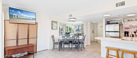 Ett modernt kök med en köksö i mitten, en matplats med bord och stolar samt en tv-skärm som visar programmet ”hosteeva”.