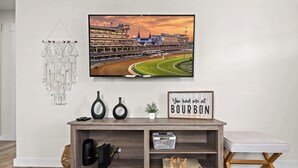 A living room with a television displaying a racing track, a macrame wall hanging, and a sign that reads "You had me at BOURBON."