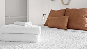 A neatly made bed with white bedding and brown pillows, a stack of white towels, and a wall-mounted light fixture.