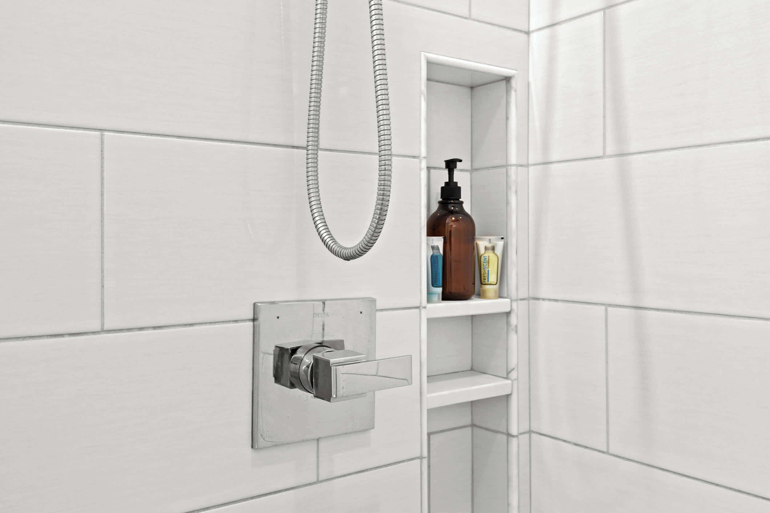 Un baño con azulejos blancos, un cabezal de ducha y un estante con artículos de aseo.