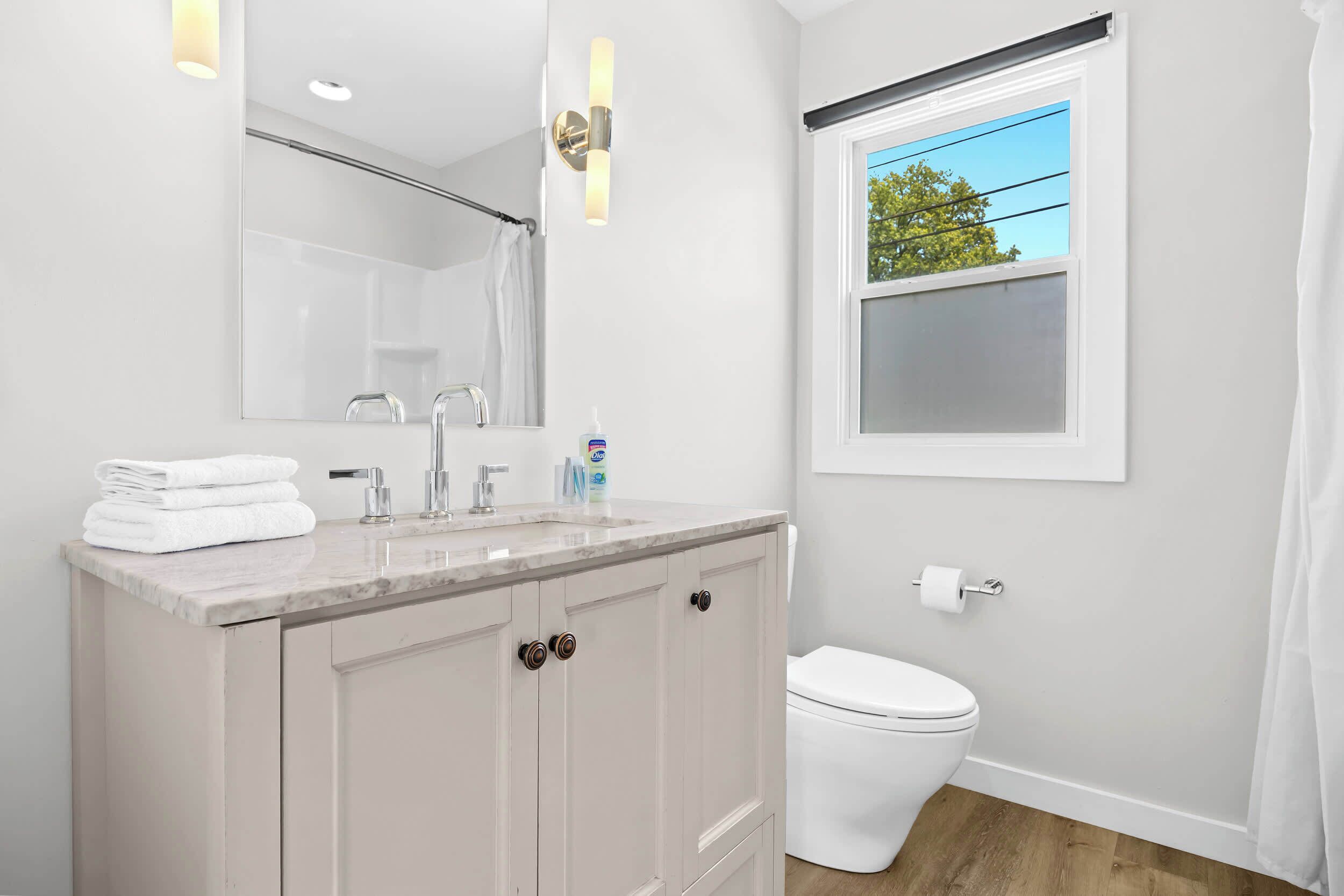 A modern bathroom with a marble sink, a large mirror, a toilet, and a window with a view of trees.
