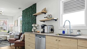 A modern kitchen with a dishwasher, a coffee maker, and a sink. There is a green wall decoration and a window with blinds.