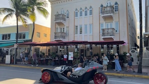 A street scene with a vintage car parked in front of a building with balconies and a red awning.