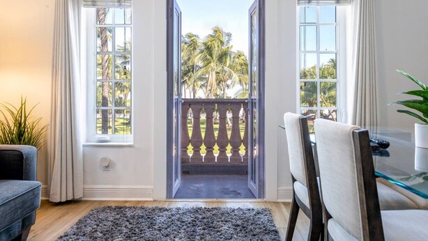 A room with a view of palm trees through a glass door, a dining area with a table and chairs, and a grey rug on the floor.