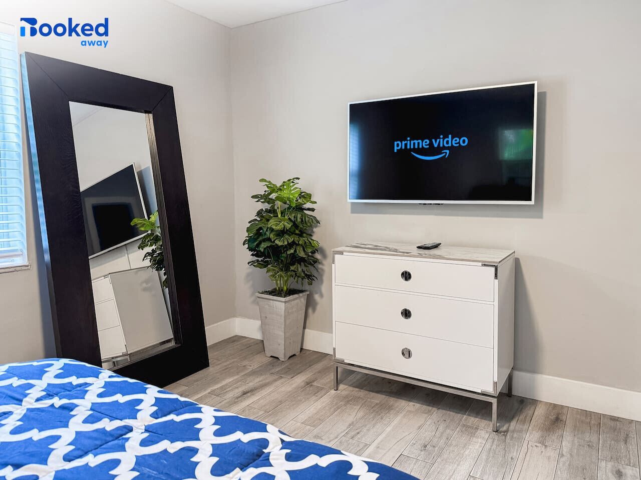 A hotel room with a bed, a TV displaying 'prime video', a dresser, a mirror, and a potted plant.