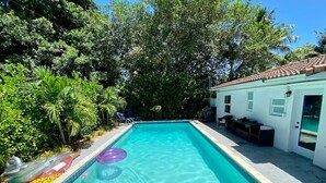 Una piscina rectangular con agua azul cristalina, rodeada de abundante vegetación y un edificio blanco con techo de tejas.