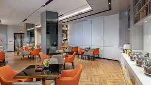 Un comedor moderno con sillas y mesas naranjas, una cocina al fondo y un mostrador de buffet con recipientes de comida.