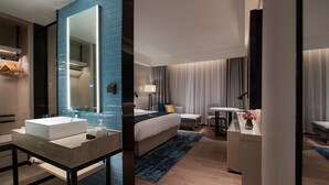 Una habitación de hotel moderna con un espejo grande, un lavabo blanco, una cama con un tapete azul y un escritorio con una lámpara.