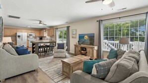 A spacious living room with a large sofa, a coffee table, and a TV. There is a kitchen area with wooden cabinets and a dining table. The room has large windows with blinds and a ceiling fan. - 'A Shore Thing' - Pool - North Myrtle Beach! (North Myrtle Beach)