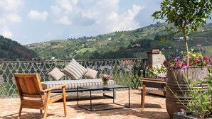 A terrace with a wooden bench, a striped sofa, and a table. There are potted plants with flowers. The terrace overlooks a hilly landscape. - Luxury Langhe Pool Vigne Di Fagnano (Santo Stefano Belbo)