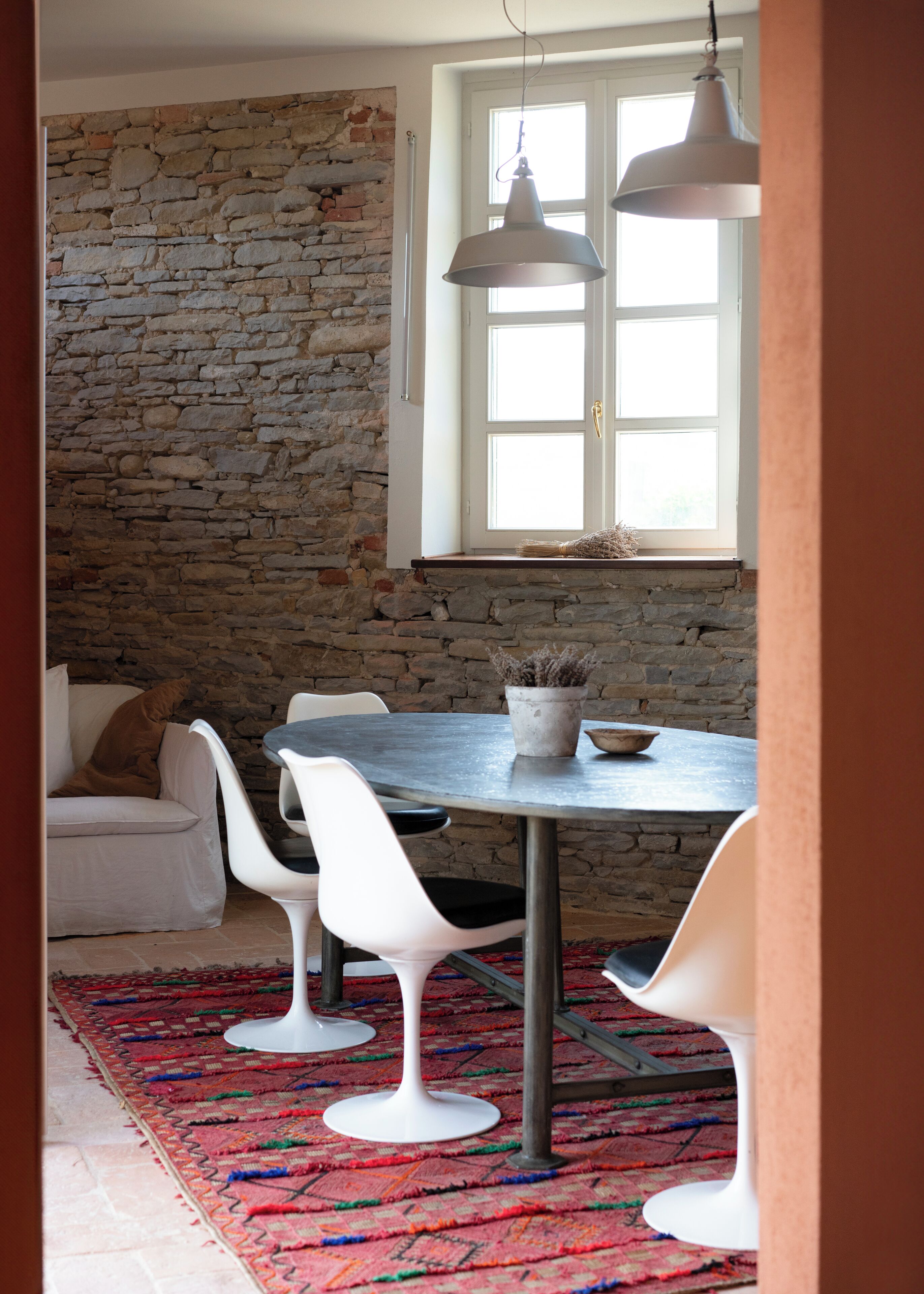 Un espace repas avec un mur en pierre, des chaises blanches, une table ronde et des lampes suspendues.