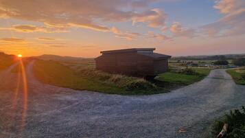 A gravel path leading to a wooden cabin with a sloped roof, surrounded by grassy fields and a sunset in the background.