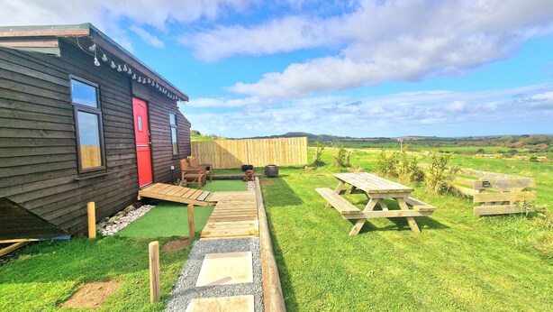 A wooden cabin with a red door, a small deck, and picnic tables in a grassy area with a view of rolling hills. - Sea View hut Sleeps 4/6 (Holyhead)