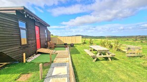 A wooden cabin with a red door, a small deck, and picnic tables in a grassy area with a view of rolling hills. - Sea View hut Sleeps 4/6 (Holyhead)