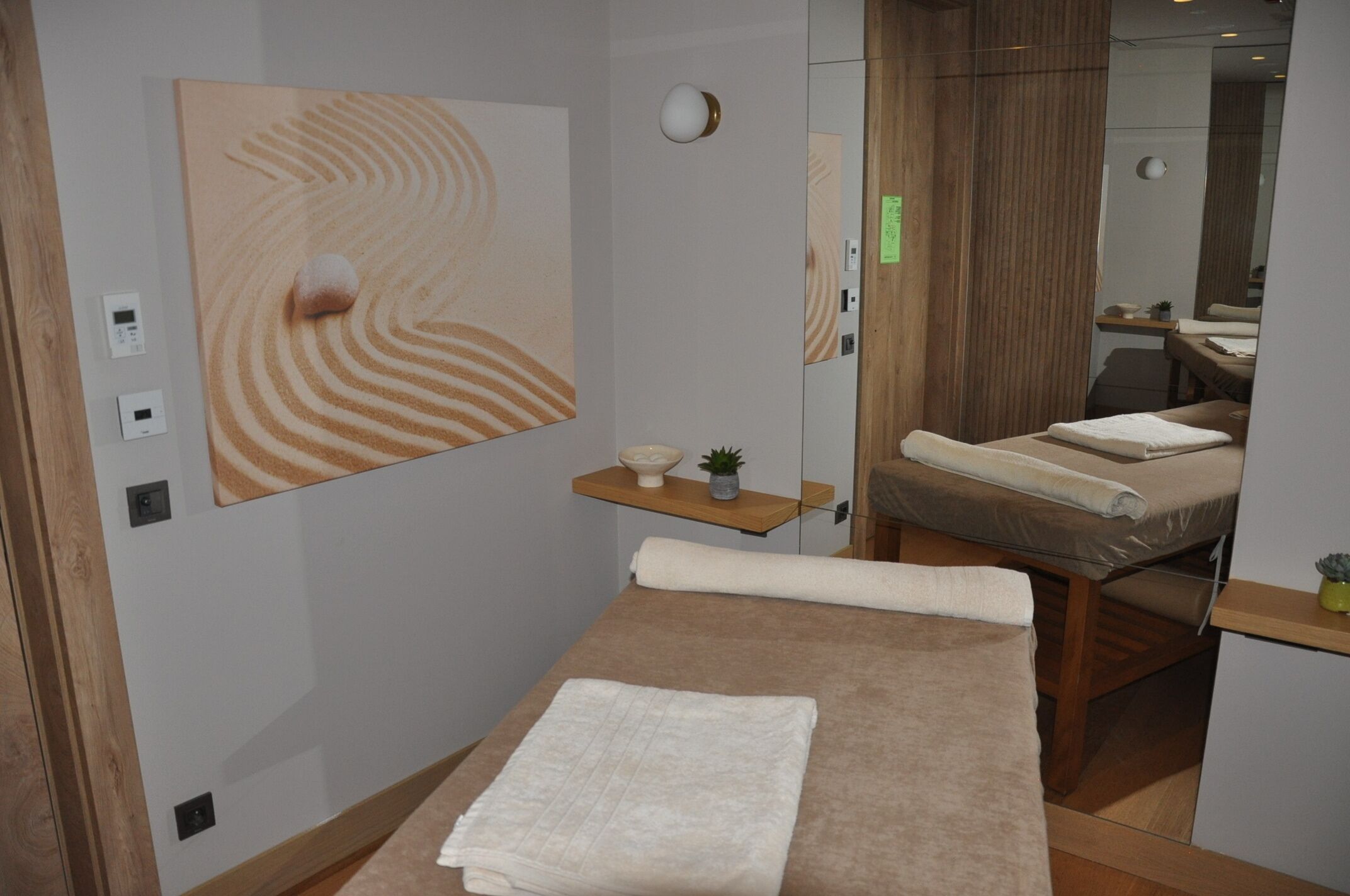 Un moderno cuarto de spa con una camilla de masaje, un cuadro abstracto grande y un estante de madera con una planta pequeña.