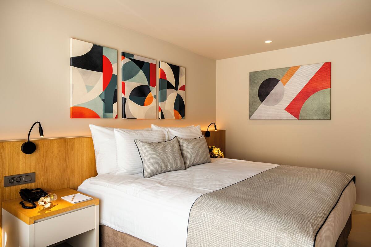 A modern hotel room with a large bed, a wooden nightstand, a desk with a lamp, and abstract wall art.