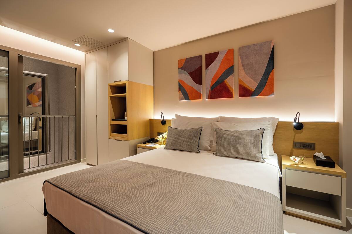 A modern hotel room with a large bed, a bedside table with a lamp, a nightstand, and abstract wall art.