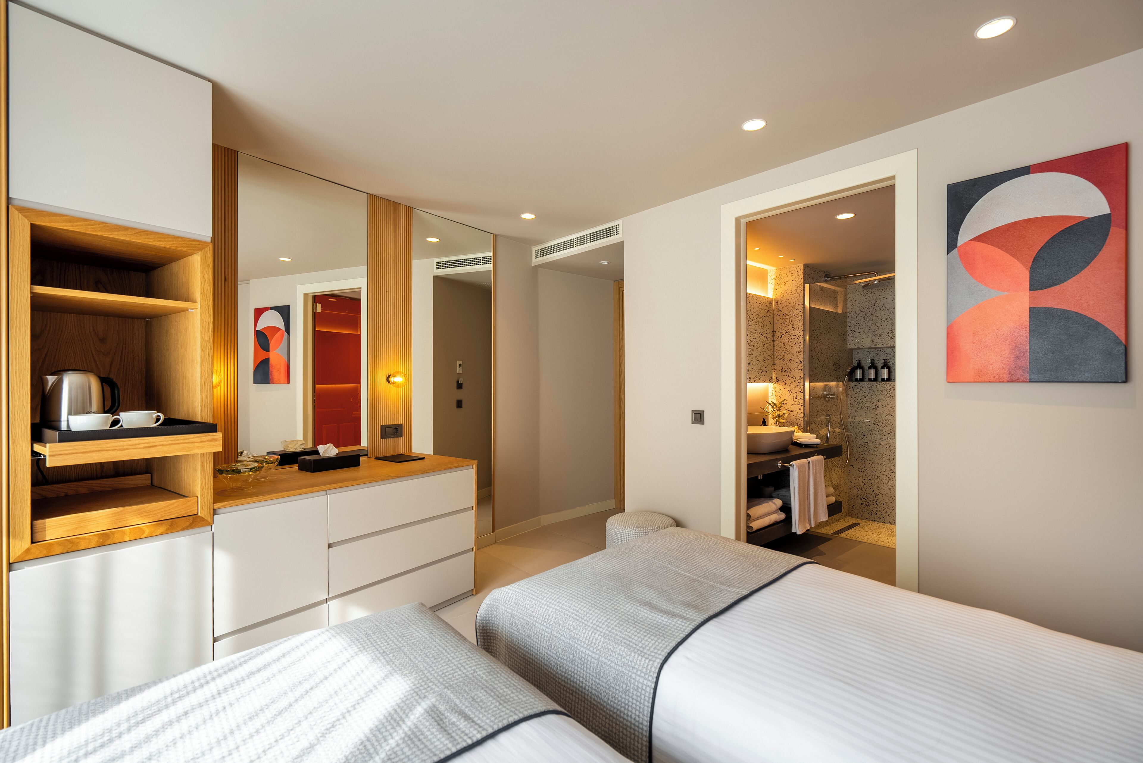 A modern hotel room with a large bed, a wooden sideboard, a built-in wardrobe, a bathroom with a shower, and a wall-mounted artwork.