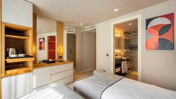 A modern hotel room with a large bed, a wooden sideboard, a built-in wardrobe, a bathroom with a shower, and a wall-mounted artwork.