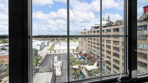 Vista a través de una ventana que muestra un edificio de varios pisos, un estacionamiento y diversas construcciones, incluyendo un tanque cilíndrico grande y un modelo de avión.
