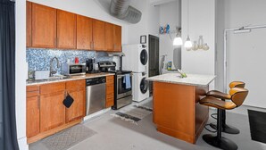 Cocina compacta con armarios de madera, microondas integrado, estufa y lavadora. Hay una pequeña isla con taburetes de bar y un lavamanos.