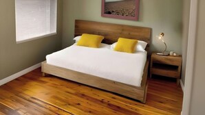 Une chambre avec un lit en bois, deux oreillers jaunes, une table de chevet avec une lampe, un ventilateur de plafond et un tableau encadré accroché au mur.