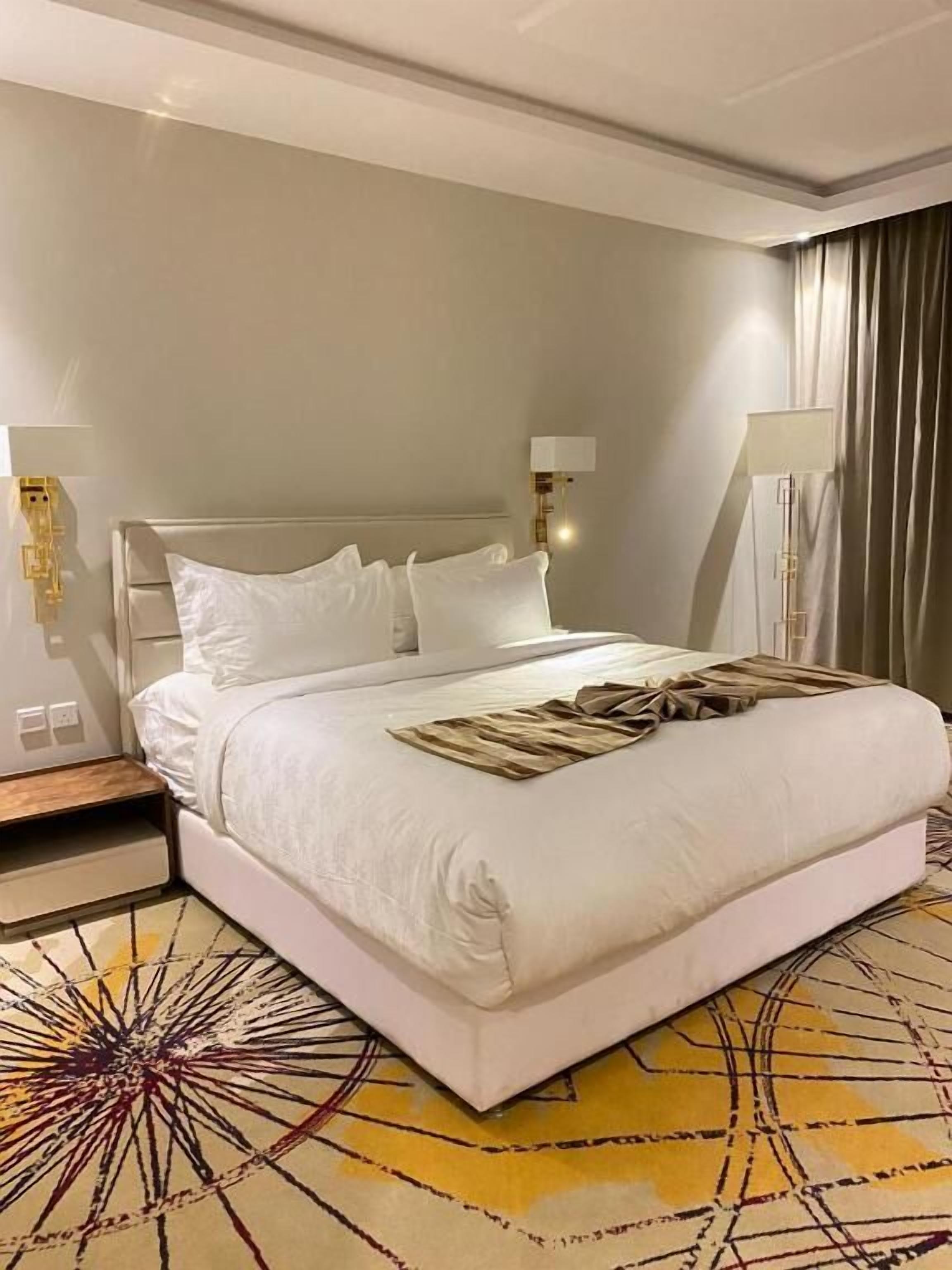 A hotel room with a large bed, a bedside table, a lamp, and a patterned rug.
