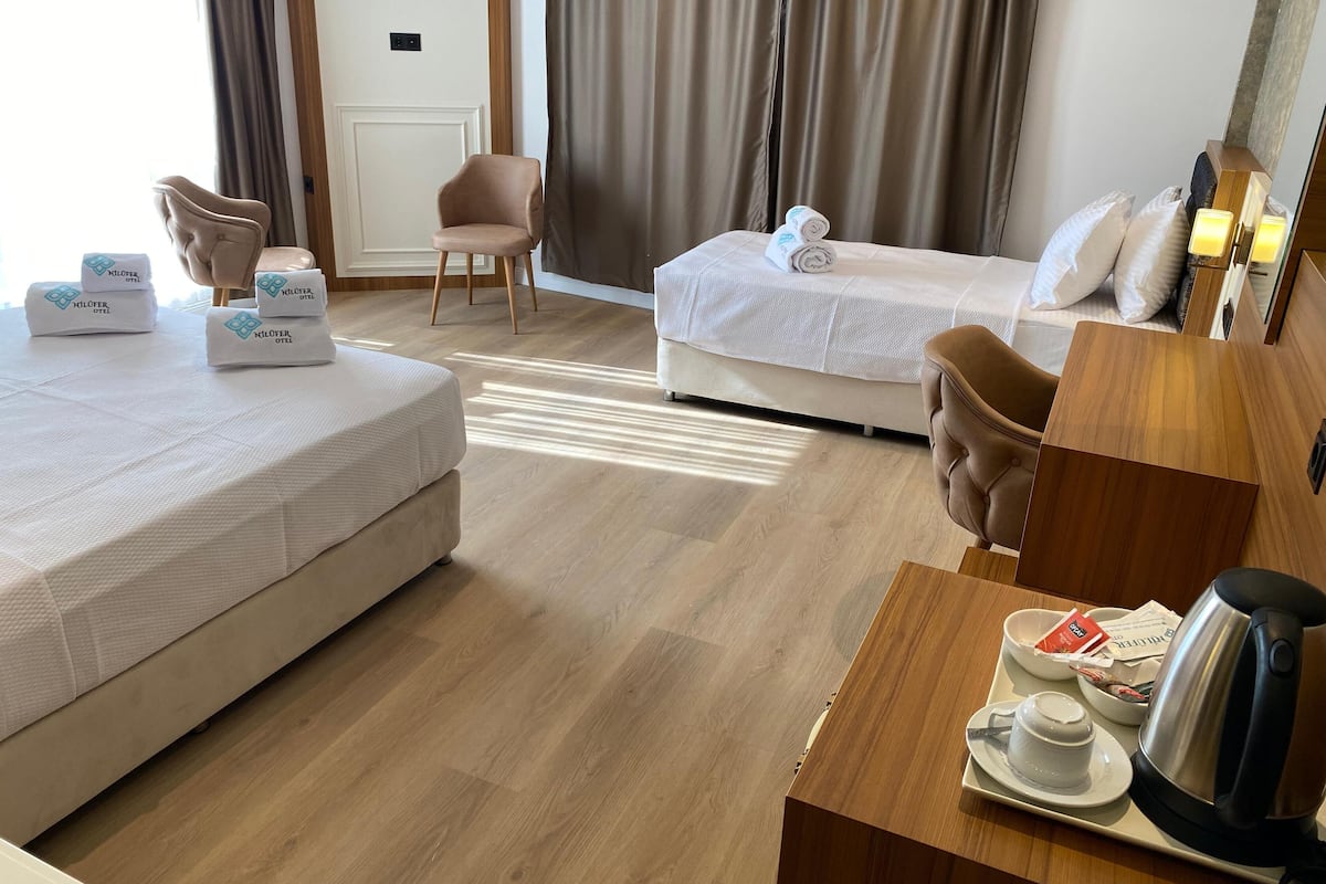 A hotel room with two beds, a wooden desk with a kettle, a chair, and a small table with a cup and saucer.