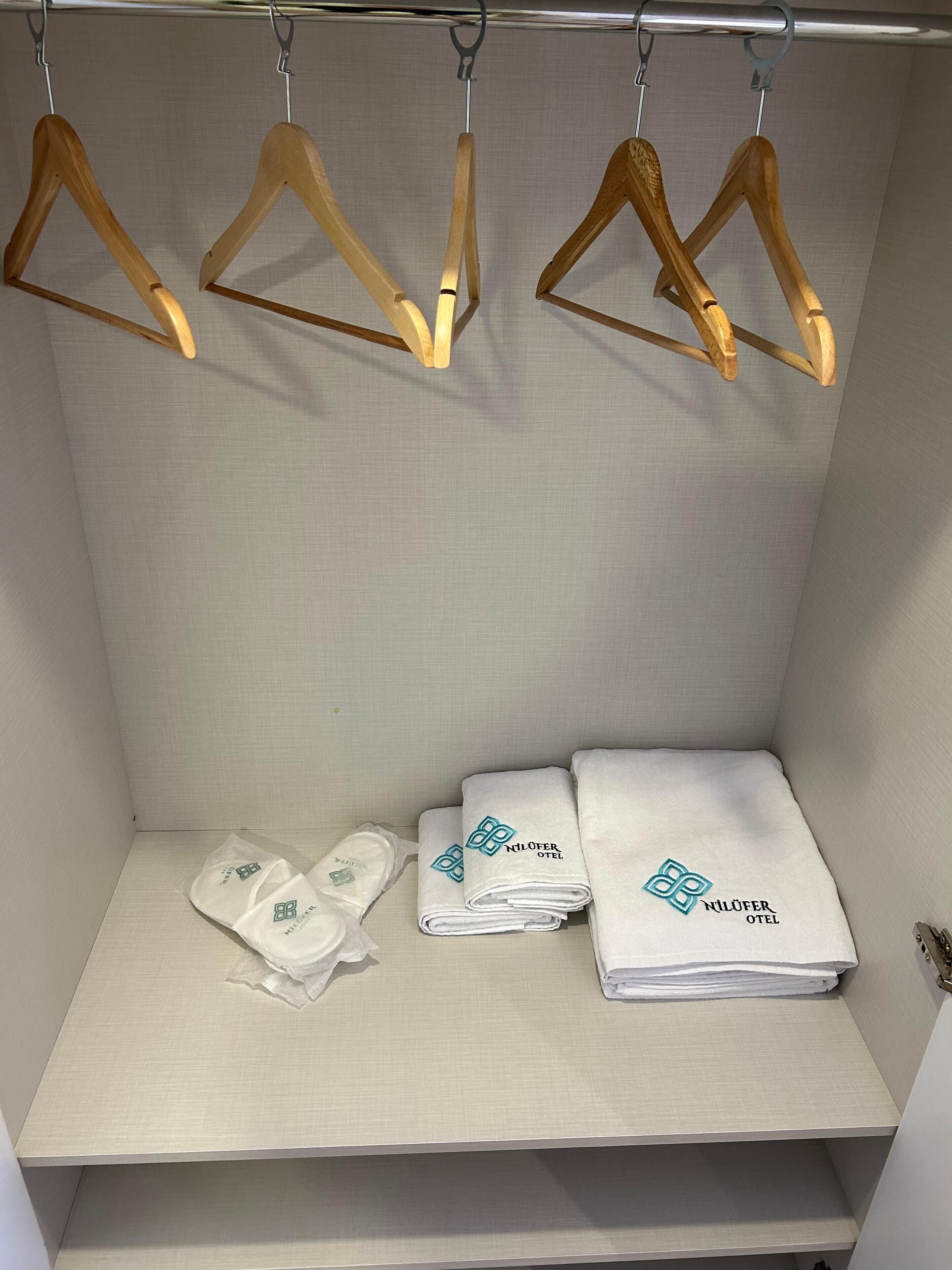 Una habitación de hotel con un estante ordenado que contiene toallas dobladas y zapatillas, con perchas de madera colgando encima.